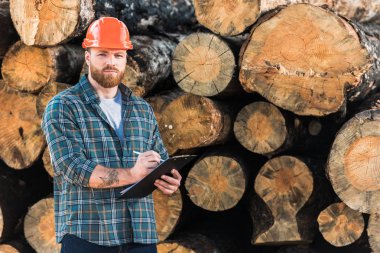lumberjack with tattooed hand in protective helmet writing in clipboard on logs background  clipart
