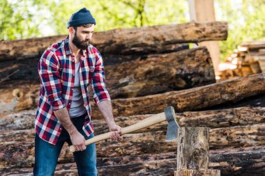 bearded lumberjack in checkered shirt chopping log at sawmill  clipart