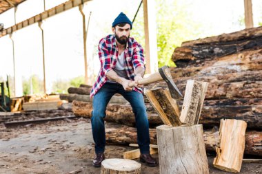 front view of bearded lumberjack in checkered shirt chopping log at sawmill  clipart