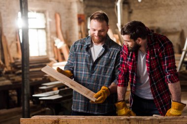 happy carpenter in protective gloves showing wooden plank to partner at sawmill clipart