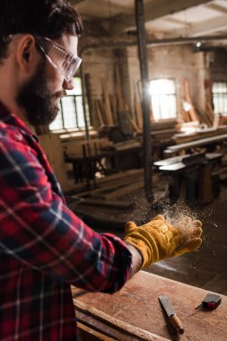 bearded craftsman in protective googles and gloves shaking off wooden chips from hands at sawmill