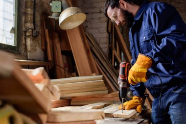 bearded craftsman in protective gloves and googles drilling wooden plank at carpentry  clipart