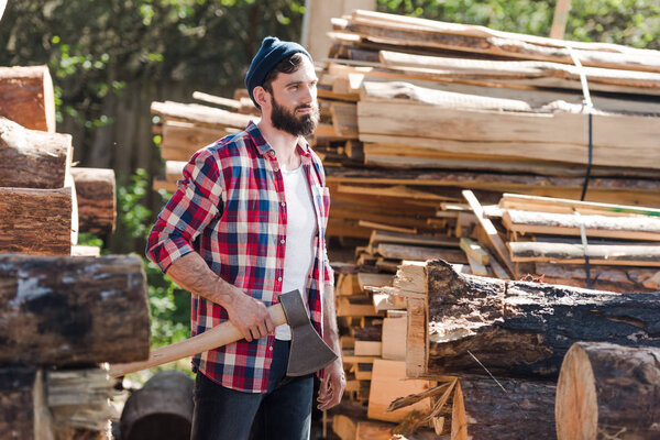 bearded lumberjack in checkered shirt standing with axe at sawmill 