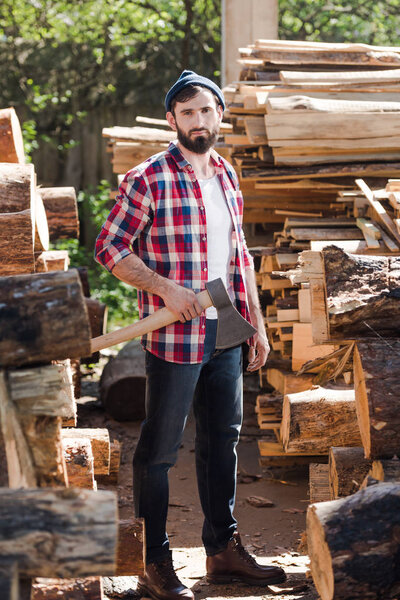 lumberjack in checkered shirt standing with axe between logs at sawmill 