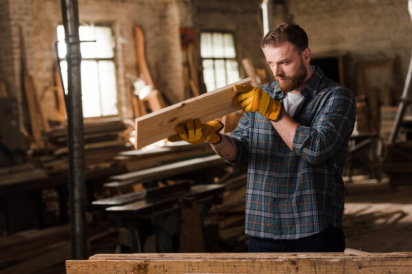 бородатый плотник в защитных перчатках проверяет деревянную доску на лесопилке
