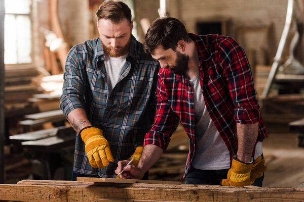  bearded carpenter in protective gloves pointing on wooden plank to partner with pen in hand 