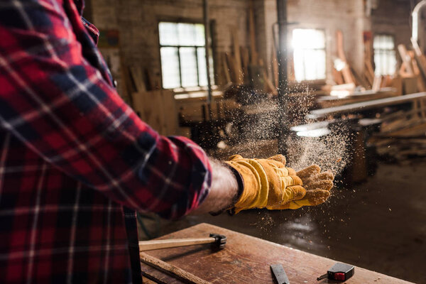 cropped shot of craftsman in protective gloves shaking off wooden chips from hands at sawmill