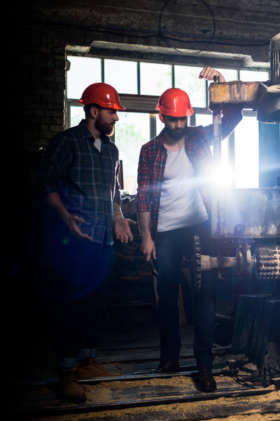 two bearded workers in protective helmets using machine tool at sawmill 