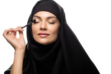 tesettürlü genç Müslüman kadın kirpiklerine rimel sürüyor. Gözleri kapalı beyaz tenli.