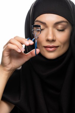 young Muslim woman in hijab using eyelash curler with closed eyes isolated on white clipart