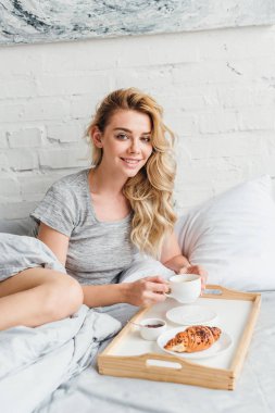 happy woman holding cup of coffee near wooden tray with croissant on bed  clipart