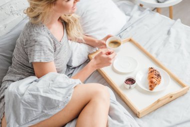 cropped view of cheerful woman holding cup of coffee near wooden tray with croissant on bed  clipart