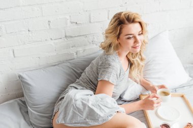 beautiful woman holding cup of coffee near wooden tray on bed  clipart