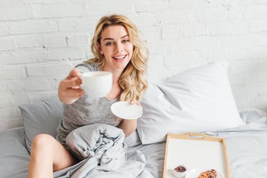 Yatakta elinde fincan kahve ve çay tabağı tutan mutlu kadının seçici odağı 