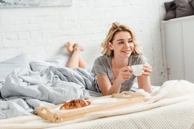 selective focus of happy girl holding cup of coffee near tray with tasty croissant on bed  clipart