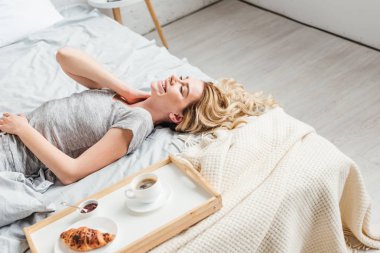 happy girl lying on bed near croissant with cup of coffee on tray  clipart