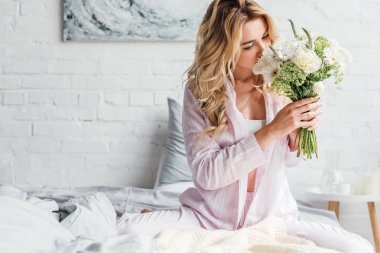 Genç bir kadın yatak odasında çiçekleri koklarken yüzünü kapatıyor. 
