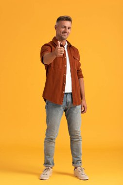 Neşeli adam turuncu üzerinde dururken baş parmağını gösteriyor. 