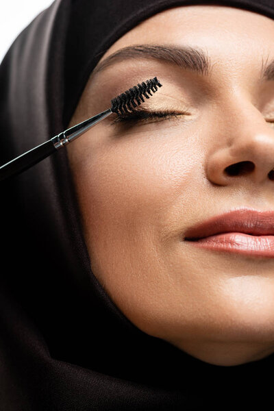 close up view of young Muslim woman in hijab applying mascara on eyelashes with closed eyes isolated on white