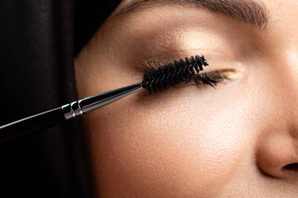 close up view of young Muslim woman in hijab applying mascara on eyelashes with closed eyes