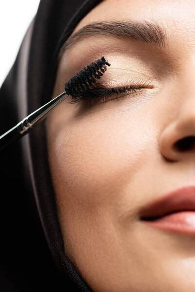close up view of young Muslim woman in hijab applying mascara on eyelashes with closed eyes isolated on white