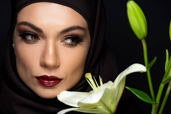 attractive Muslim woman in hijab with red lips near lily isolated on black