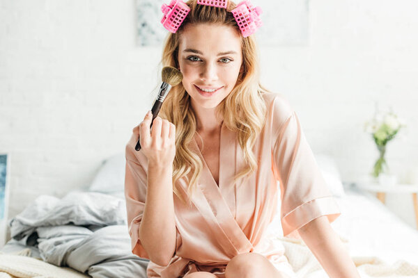 cheerful girl in hair curlers holding cosmetic brush near face 