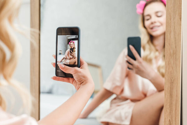 selective focus of smiling woman with hair curlers holding smartphone while taking photo near mirror 