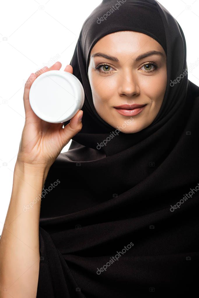 smiling young Muslim woman in hijab holding container with cosmetic cream isolated on white