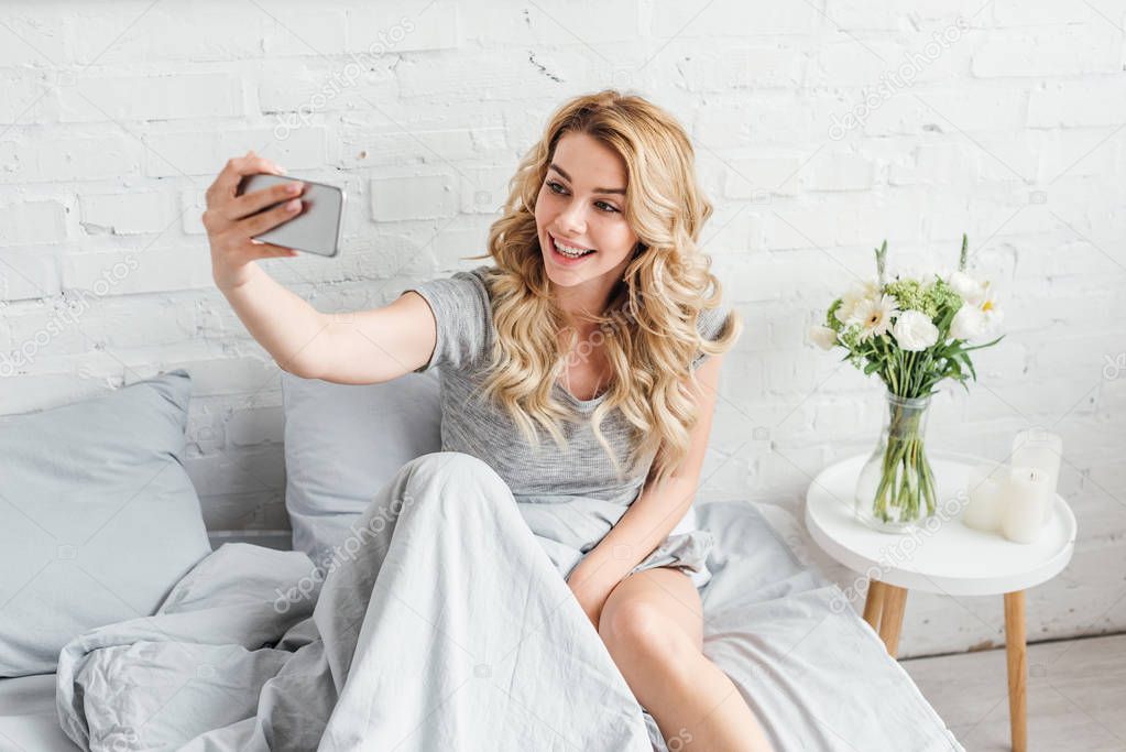 selective focus of happy girl taking selfie near vase with flowers on bedside table 