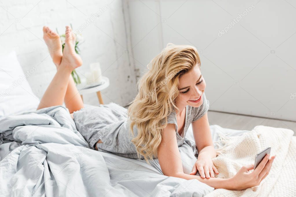 selective focus of smiling girl using smartphone while lying on bed 