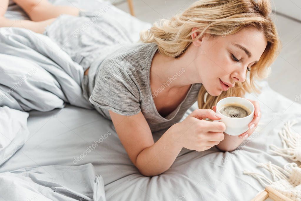 selective focus of attractive girl looking at cup with coffee while lying on bed 
