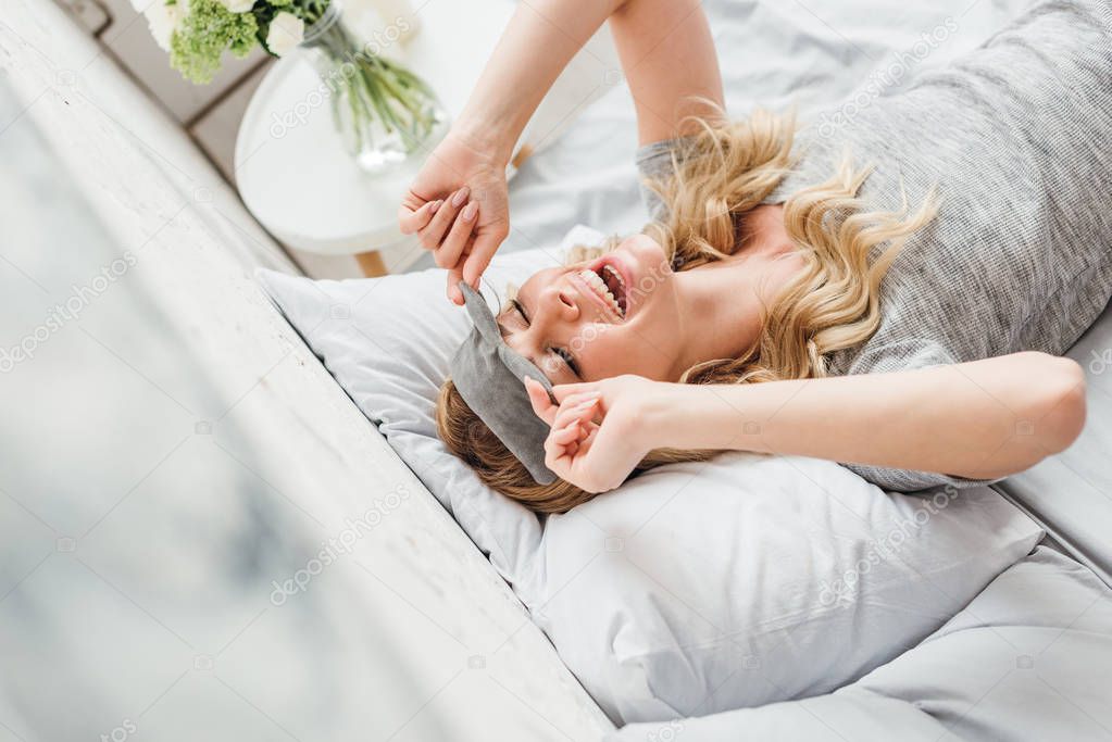 selective focus of cheerful girl touching sleeping mask in bedroom 