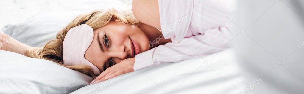 panoramic shot of cheerful woman with eye mask lying on bed at home 