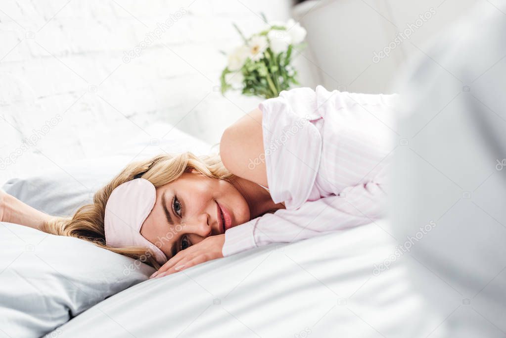 selective focus of smiling woman with eye mask lying on bed at home 