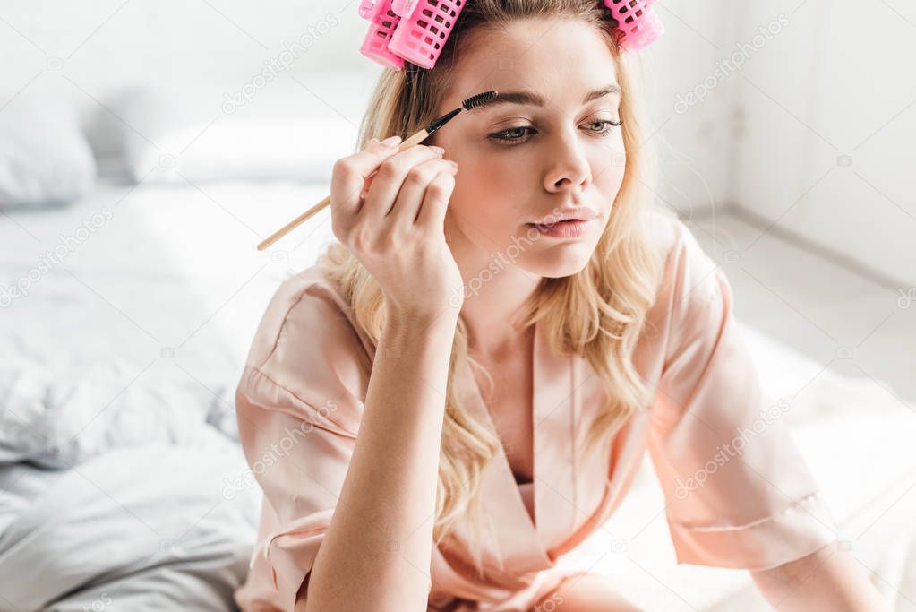 beautiful woman in pink hair curlers styling eyebrows in bedroom 