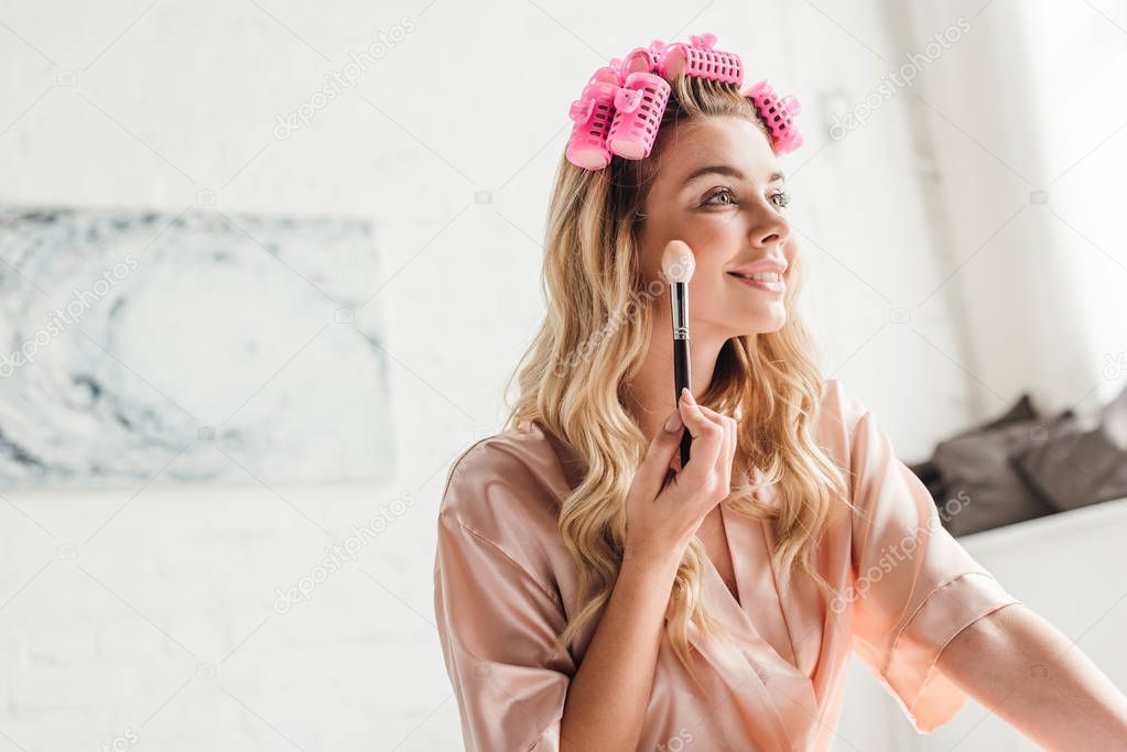 cheerful woman in pink hair curlers holding cosmetic brush near face in bedroom 