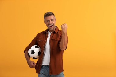 happy man standing with football and celebrating isolated on orange  clipart