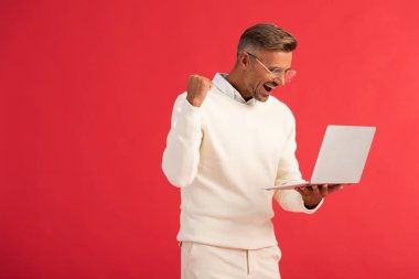 excited and stylish man in glasses holding laptop isolated on red  clipart