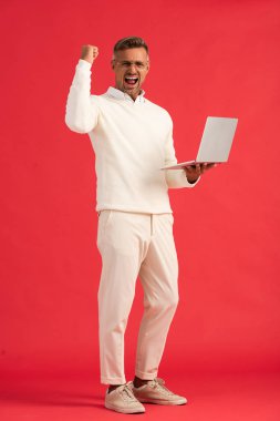 excited and stylish man in glasses holding laptop on red  clipart