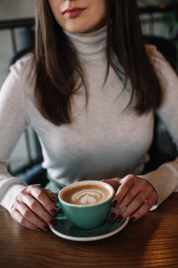 Kafenin balkonunda kapuçinolu ahşap masada oturan genç bir kadın görüntüsü.