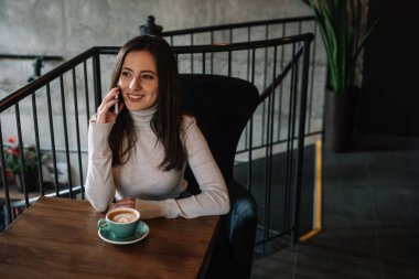 Kapuçino ile ahşap masada oturan ve kahve dükkanının balkonunda akıllı telefondan konuşan gülümseyen genç kadın.