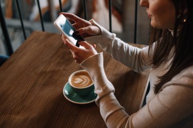 Kapuçinolu ahşap masada oturan genç bir kadının kahve dükkanının balkonunda akıllı telefon kullanması.