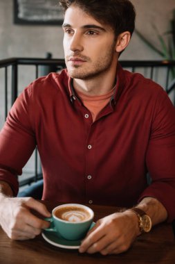 serous young man sitting at wooden table with cappuccino in coffee shop clipart