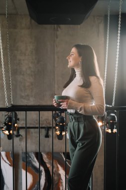 Kahvecide kapuçinoyla balkonda duran gülümseyen kadının yan görüntüsü.