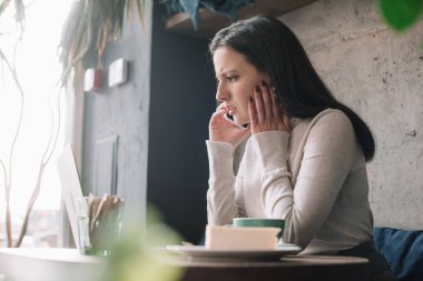 Yeşil bitkilerin seçici odağı ve dizüstü bilgisayar kullanan ve kahve dükkanında akıllı telefondan konuşan üzgün serbest çalışan.