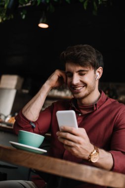 Kablosuz kulaklıklı gülümseyen adam masada elinde kahve ile akıllı telefon kullanıyor.