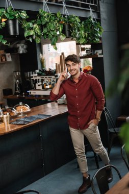 smiling handsome man walking near bar counter and talking on smartphone