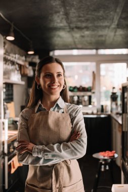 Önlüklü gülümseyen barista kollarını kavuşturarak kafede duruyor.