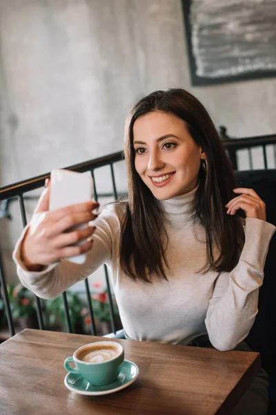 年轻女子笑着和卡布奇诺一起坐在木制桌旁 在咖啡店的阳台上用智能手机自拍 — 图库照片